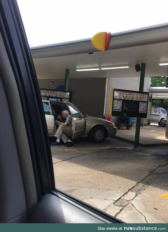 An elderly man sitting outside of his car door spoon feeding his wife ice cream
