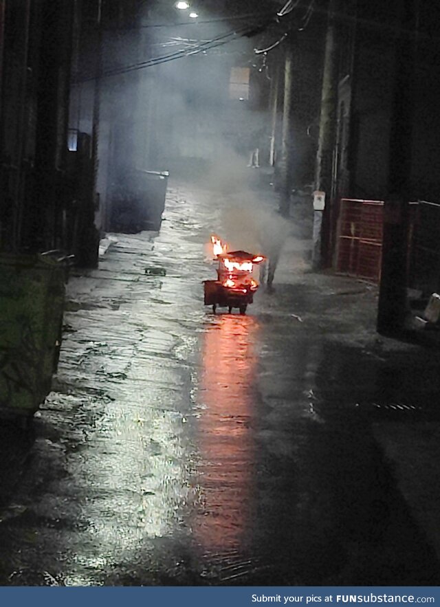 Man pushing a flaming shopping cart down an alley