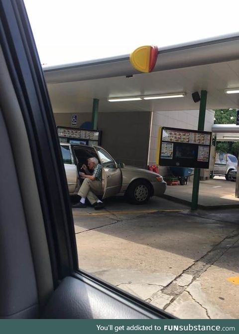 An elderly man sitting outside of his car door spoon feeding his wife ice cream